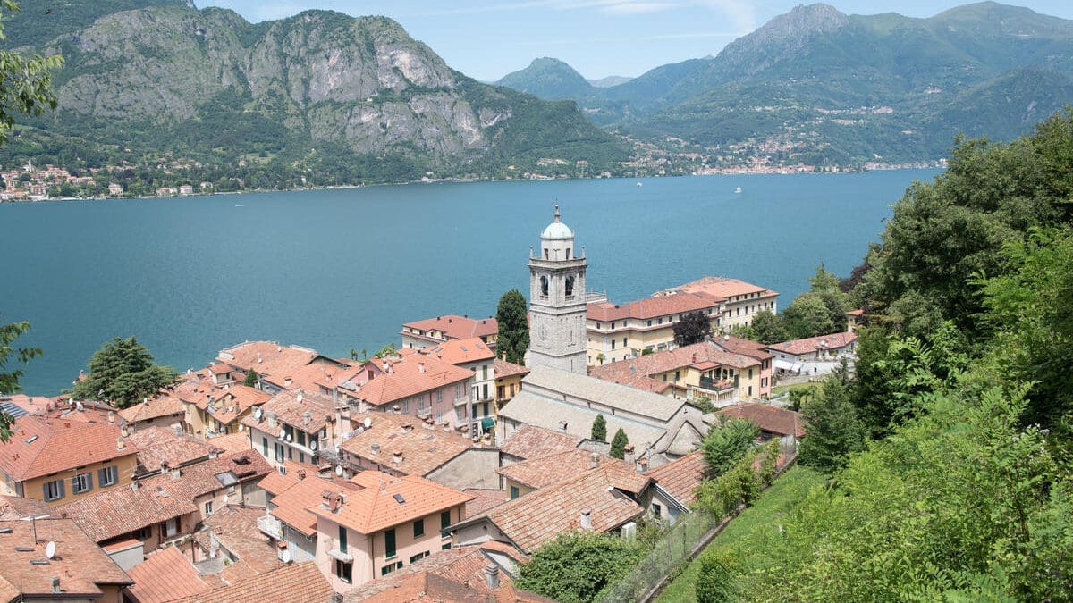 view of a lakeside town with red-roofed buildings and a central church tower, surrounded by green hills and a large blue lake