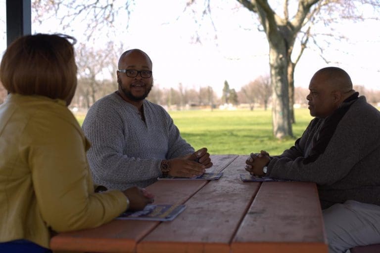 two men and a woman sitting at a picnic table looking at one another talking.