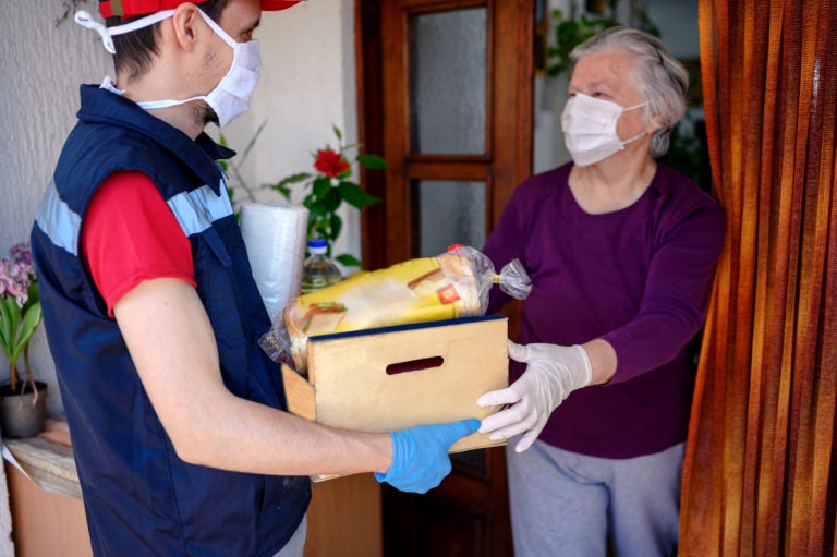 a man delivering bread in a crate of food to an elderly woman both wearing facemasks.