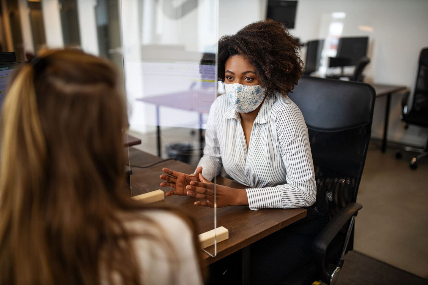 woman sitting in a chair speaking to another woman wearing a facemask.