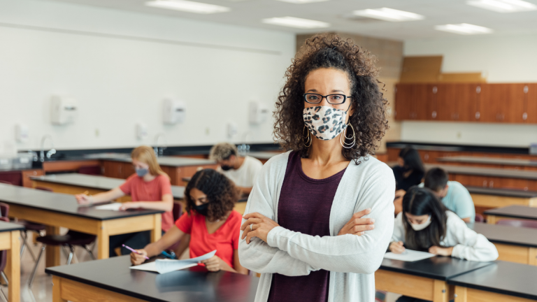 woman standing with her arms crossed wearing a facemask with men and women sitting in the background at desks taking a test.