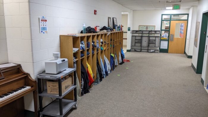 A photo of a school hallway used as a testing site, with an air sampling device on a cart on the left, nestled between an upright piano and a series of cubbyholes for children's jackets.
