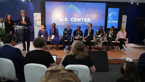 man standing at podium with people behind him sitting in chairs, speaking to a crowd of people also sitting in chairs