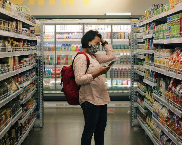 woman wearing a mask and standing in the aisle of a grocery store looking at food on a shelf