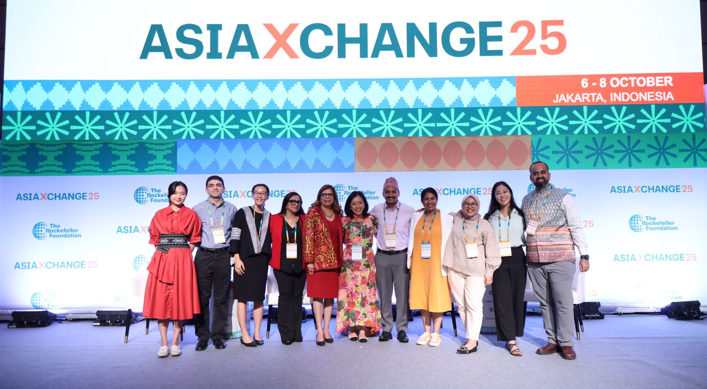 a group of eleven attendees wearing conference badges pose together on stage at AsiaXchange25 in Jakarta, dressed in colorful business attire and traditional clothing