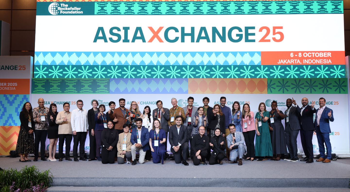 a large group of AsiaXchange25 attendees wearing conference badges pose together on stage in front of the event backdrop in Jakarta, Indonesia