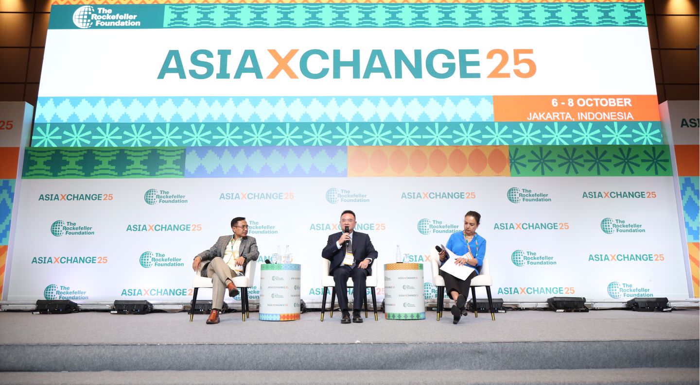 three panelists participate in a discussion on stage at AsiaXchange25, seated in white chairs with a moderator in blue holding papers and a microphone