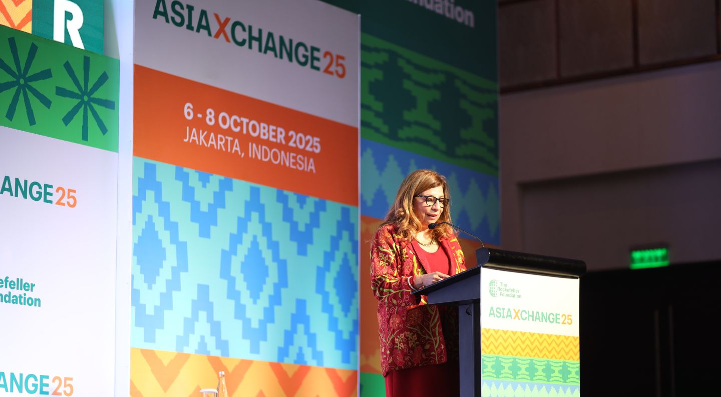 a speaker in a patterned red jacket and glasses addresses the audience from a podium at the AsiaXchange25 event in Jakarta, Indonesia
