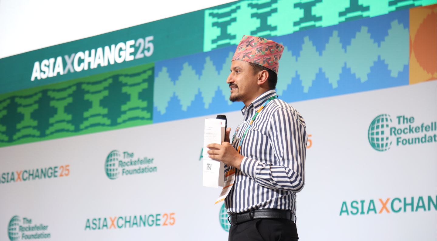a speaker wearing a traditional patterned cap and striped shirt holds a microphone while presenting at the AsiaXchange25 event