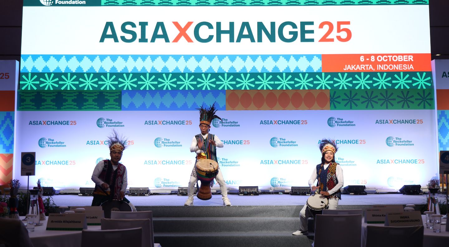 three performers in traditional Indonesian attire and feathered headdresses play drums on stage at the AsiaXchange25 event in Jakarta