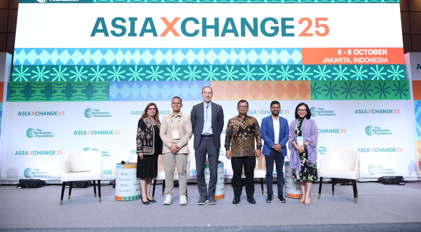 six people stand on stage at the AsiaXchange25 event in Jakarta, Indonesia, in front of a branded backdrop with colorful geometric patterns and The Rockefeller Foundation logo
