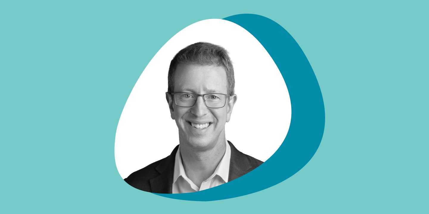 A black and white photo of a man wearing glasses who is smiling at the camera in a rounded, triangular frame, set against overlapping teal and turquoise circles on a light teal background