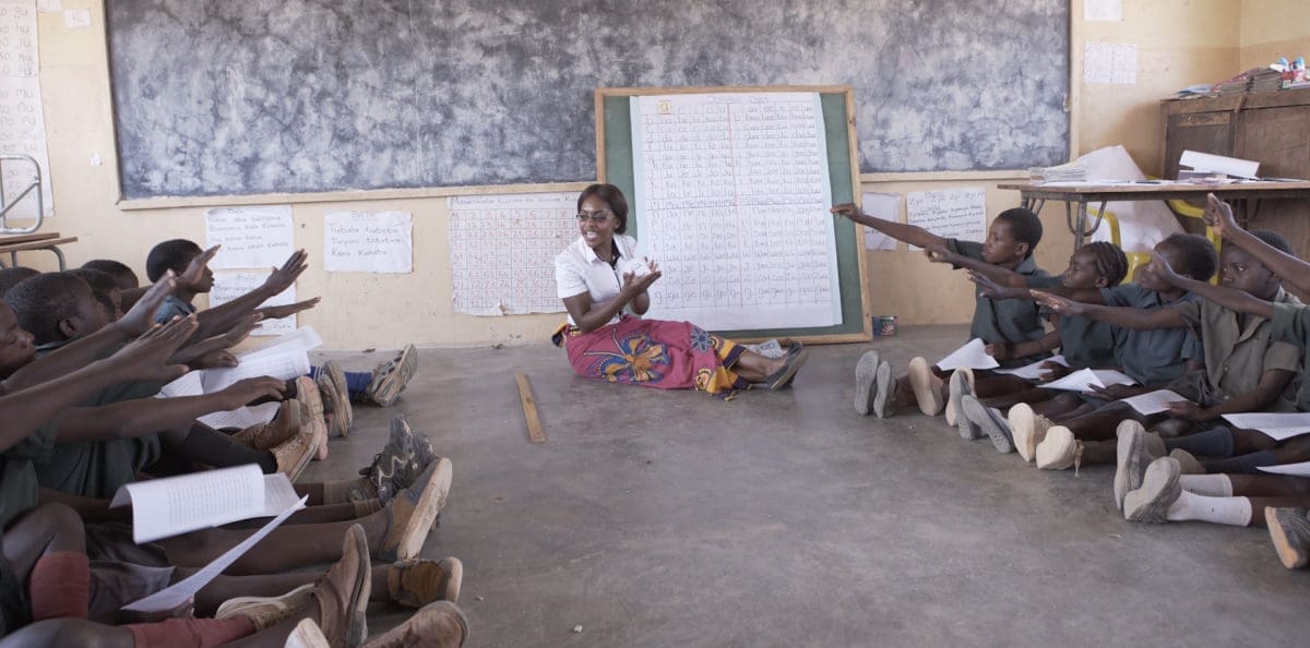 young boys and girls sitting at desks raising their hands in a classroom.
