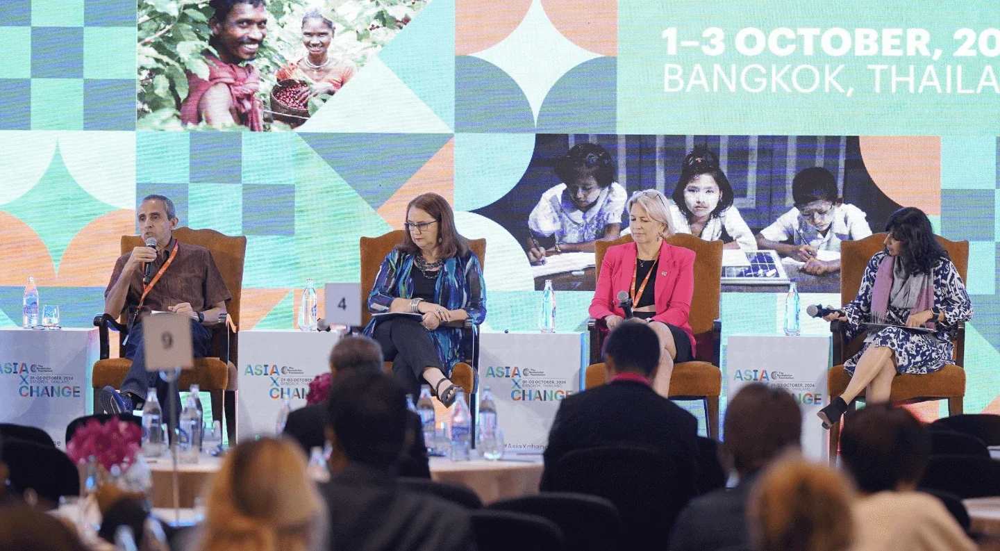 four panelists sit on stage at a conference, discussing topics related to change in Asia. The background features colorful graphics and the event date and location are displayed. Attendees are visible in the foreground, focused on the panel discussion