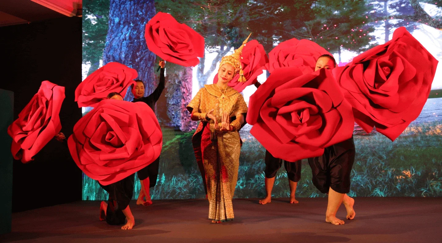 dancers perform on stage, holding large red rose props around a performer in traditional attire.
