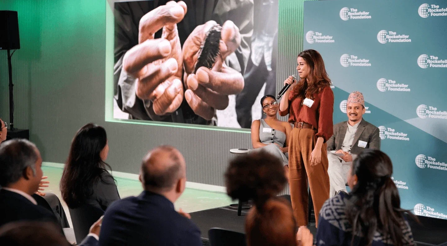 a woman speaks on stage with a microphone during a Rockefeller Foundation UNGA 2025 session, while audience members listen and a large screen behind her displays a close-up image of hands holding a small object.