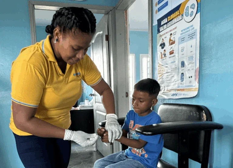 a healthcare worker wearing gloves checks a young boy’s hand in a clinic, with a health poster about safe water on the wall behind them.