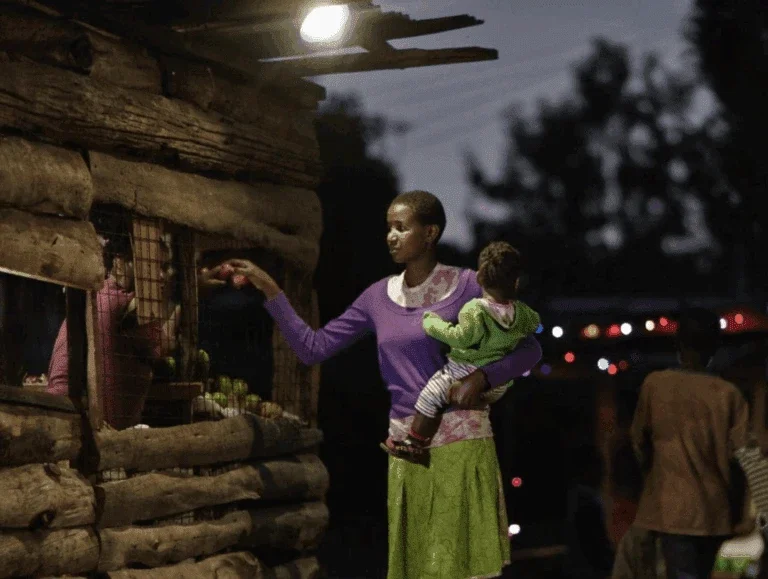 a woman holding a child buys produce at a small wooden market stall lit by an overhead solar-powered light in the evening.