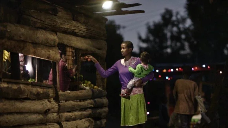 a woman holding a child buys produce at a small wooden market stall lit by an overhead solar-powered light in the evening.