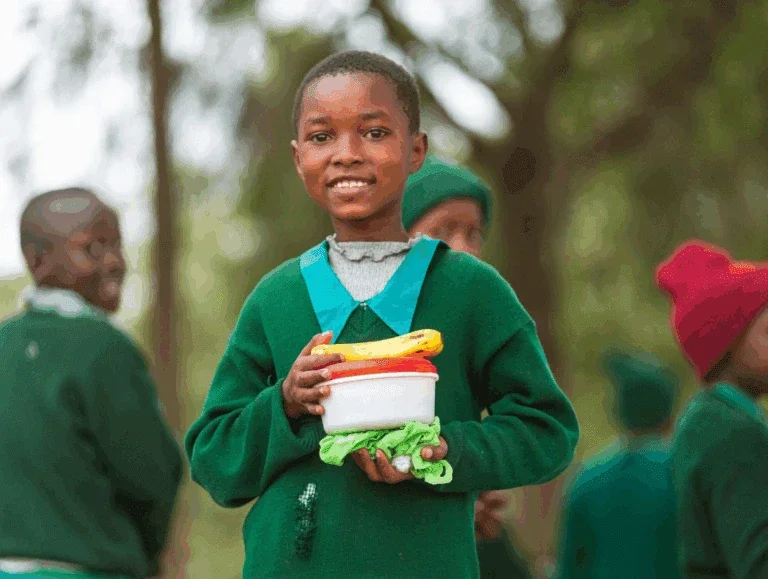 a smiling schoolchild in a green uniform holds a lunch container with a banana on top, standing outdoors with classmates in the background.