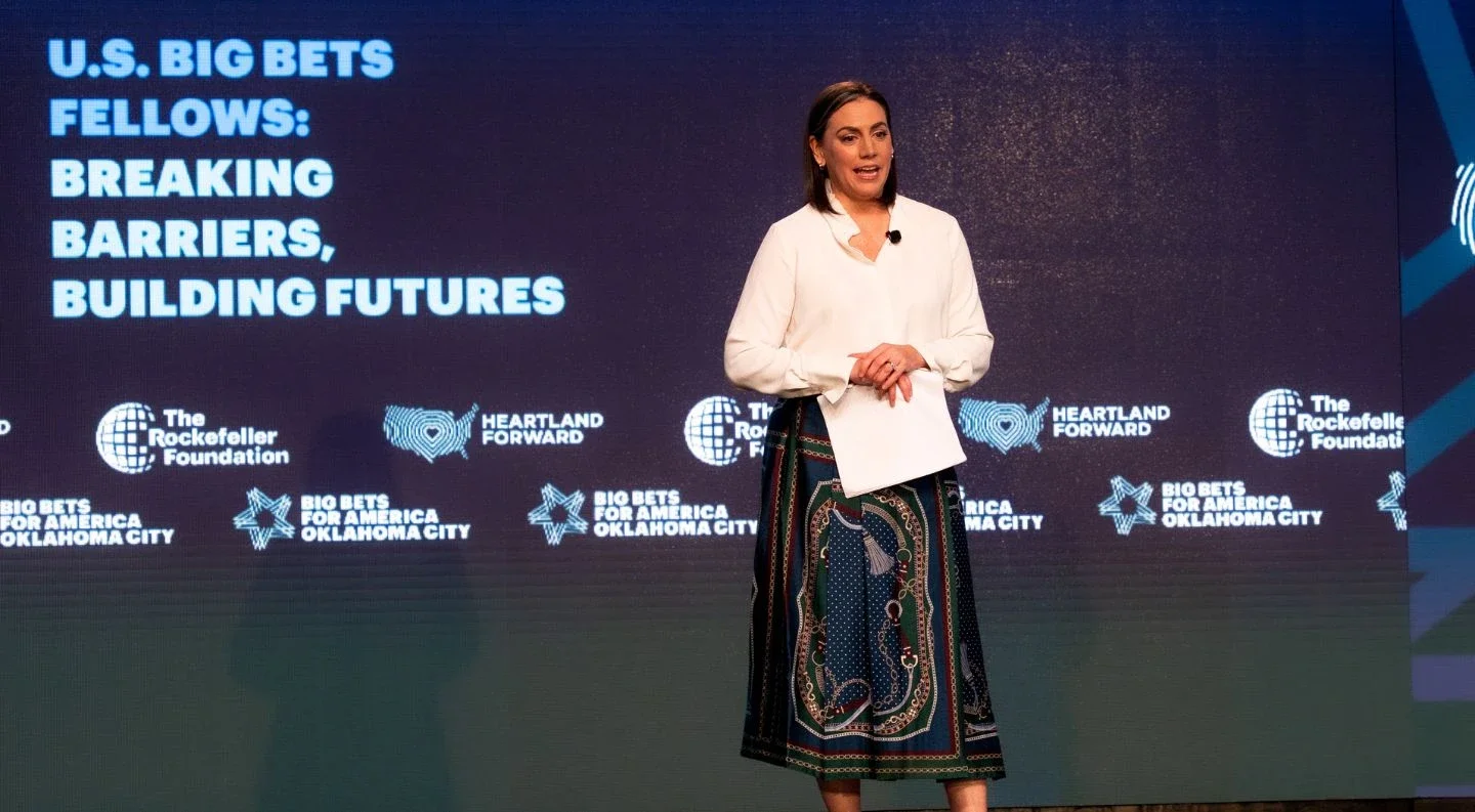 speaker presenting on stage about "U.S. Big Bets Fellows: Breaking Barriers, Building Futures" at Big Bets for America, with partner organization logos displayed on the backdrop.