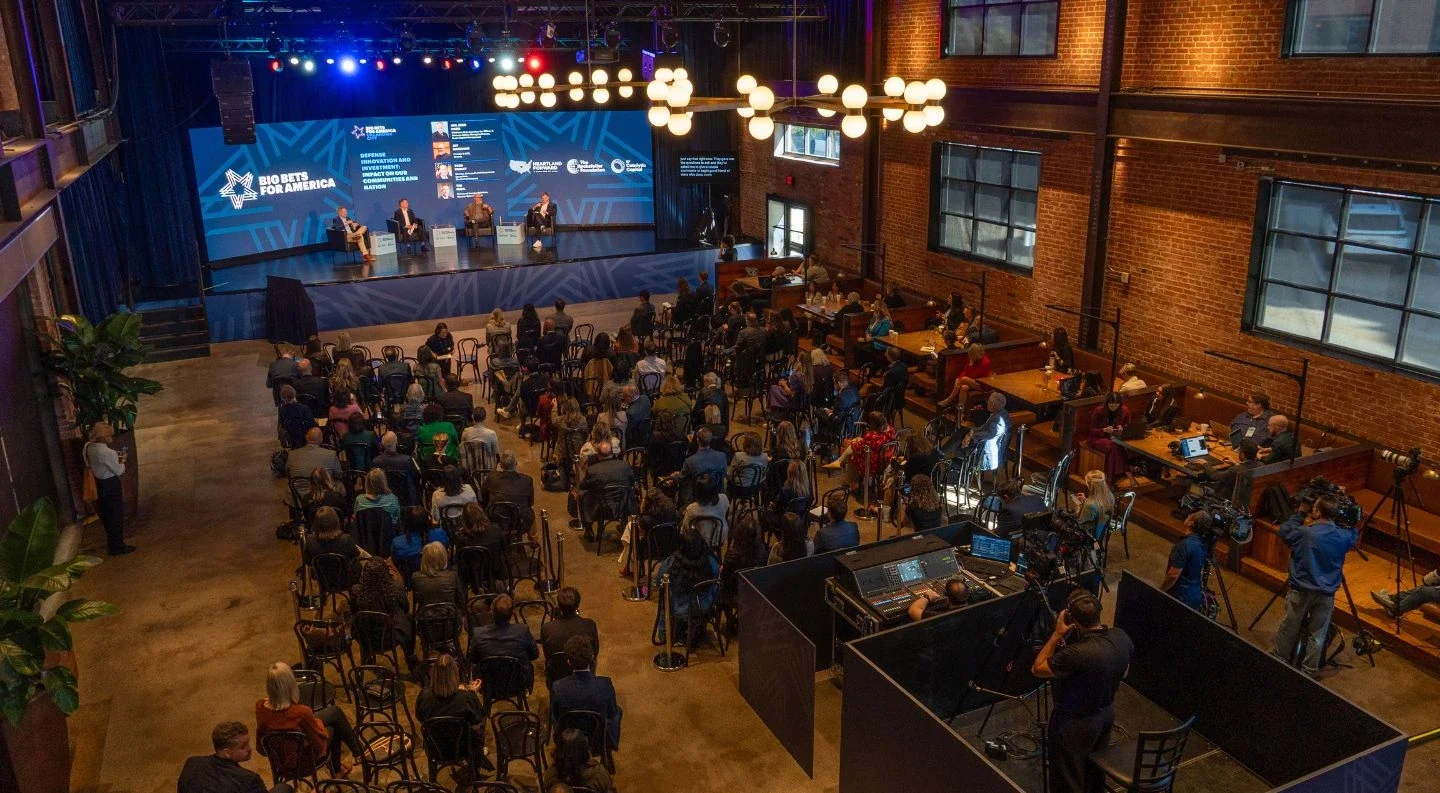 aerial view of the Big Bets for America convening in a brick-walled venue, showing attendees watching a panel discussion on stage with production equipment and café tables visible.