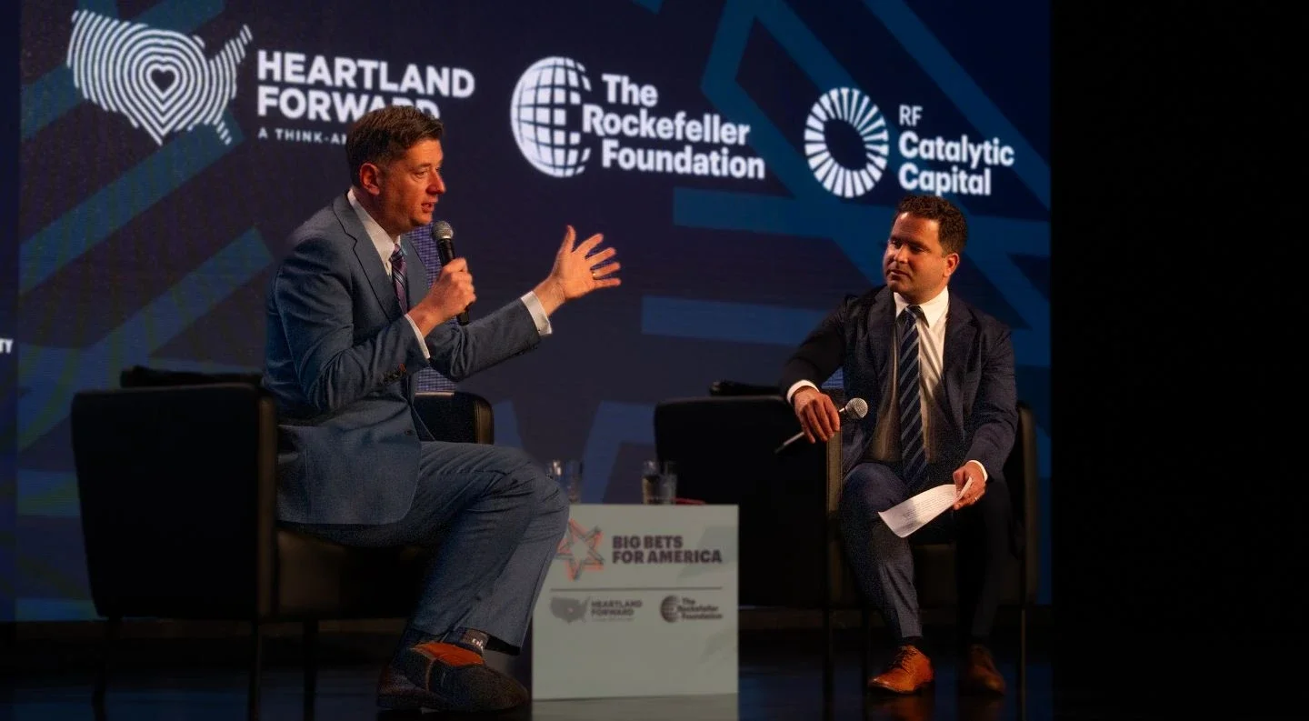 two speakers in conversation on stage at Big Bets for America, seated in chairs with event branding visible and partner logos for Heartland Forward, The Rockefeller Foundation, and RF Catalytic Capital on the backdrop.