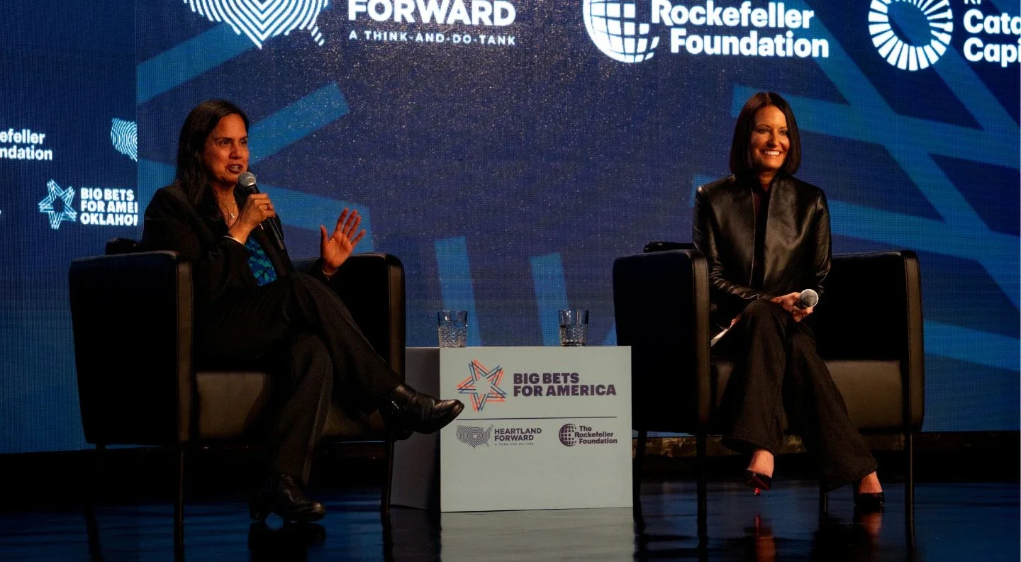 two speakers in conversation on stage at the Big Bets for America convening, seated in chairs with the event logo displayed between them and partner organization logos on the backdrop.